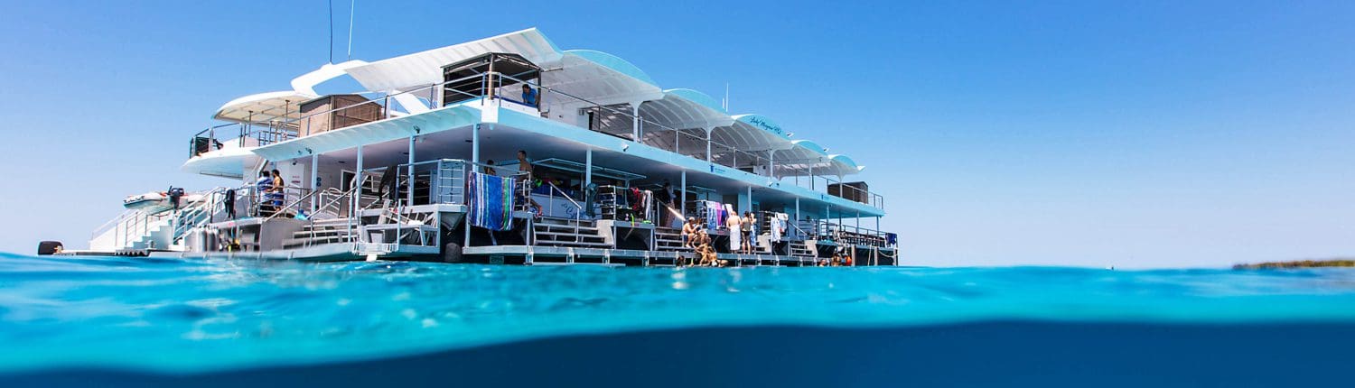 A large, multi-level floating platform from the Lady Musgrave Experience is anchored in clear blue water under a sunny sky, with part of the structure and water visible above and below the surface during one of their Great Barrier Reef Tours.