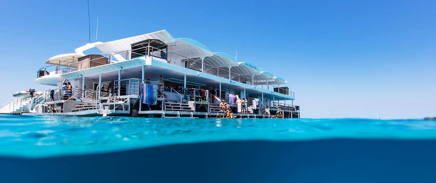 A large, multi-level floating platform from the Lady Musgrave Experience is anchored in clear blue water under a sunny sky, with part of the structure and water visible above and below the surface during one of their Great Barrier Reef Tours.