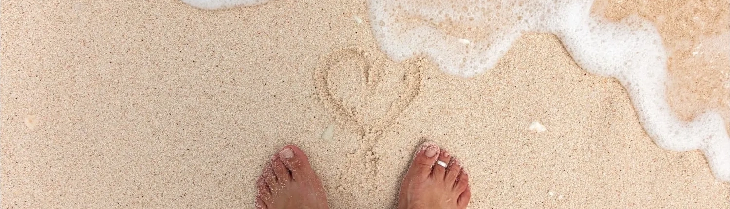 faqs-Lady-Musgrave-Experience A pair of bare feet stands on sandy beach near a heart shape drawn in the sand, as gentle ocean waves—like those found on a Lady Musgrave Experience or Great Barrier Reef tour—begin to wash over the drawing.