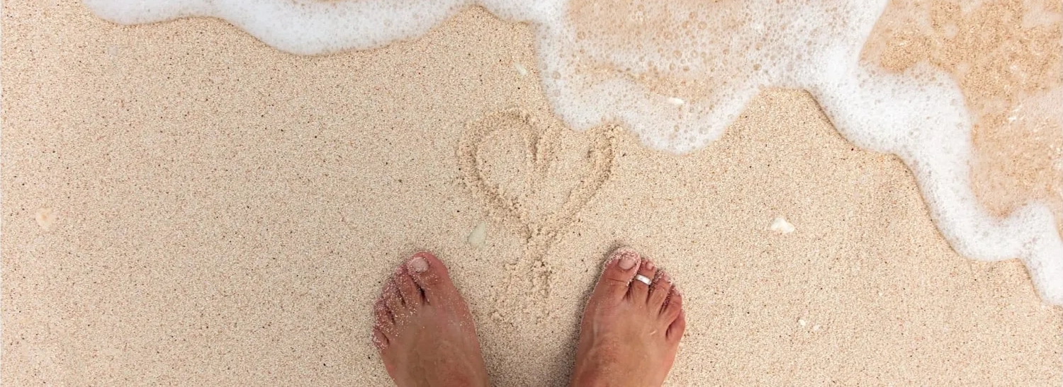 faqs-Lady-Musgrave-Experience A pair of bare feet stands on sandy beach near a heart shape drawn in the sand, as gentle ocean waves—like those found on a Lady Musgrave Experience or Great Barrier Reef tour—begin to wash over the drawing.